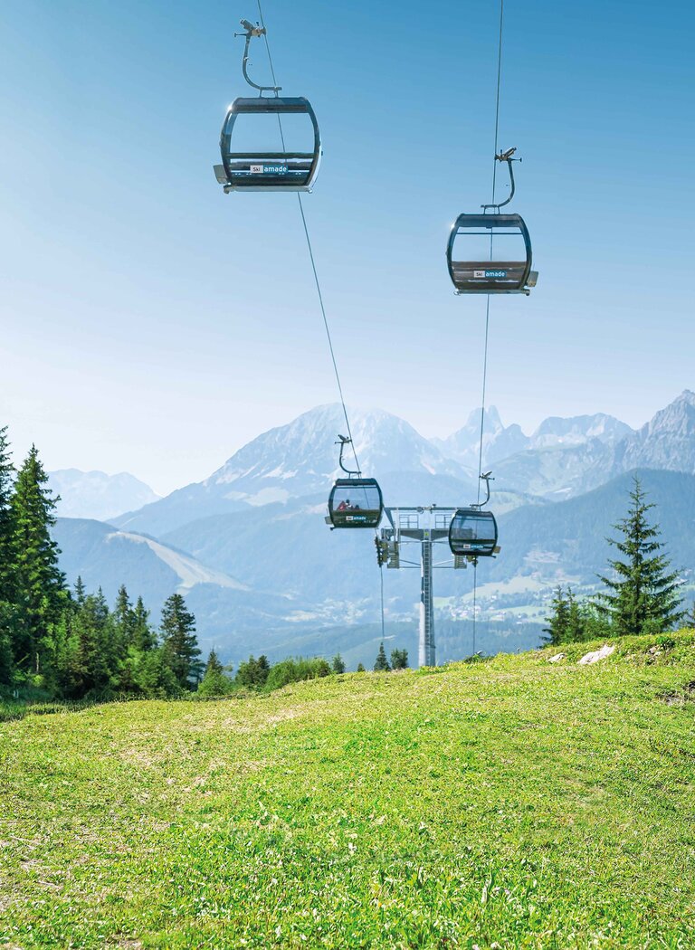 Gondeln schweben bei Sonnenschein über grüne Almwiese, im Hintergrund bewaldete Hänge und felsige Berggipfel. | © Ski amadé GmbH